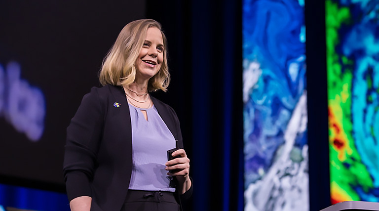 A woman stands at a podium on a stage at an Esri event, flanked by two large screens displaying images of maps